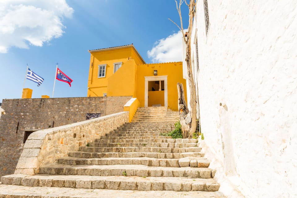 Una escalera de piedra conduce a un edificio amarillo con dos banderas, una griega y otra turca, bajo un cielo azul despejado.