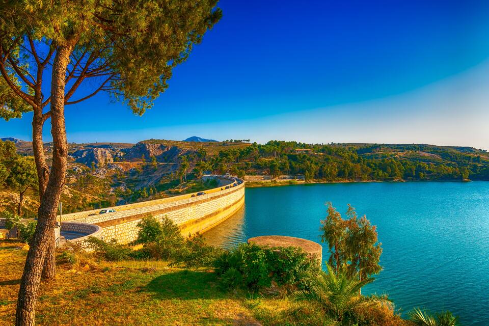 A dam with a road running alongside it, a clear blue lake, and surrounding hills.