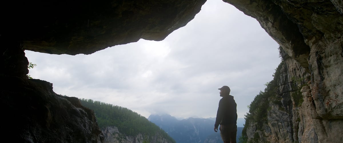A person standing in a cave opening with a mountainous landscape in the background.