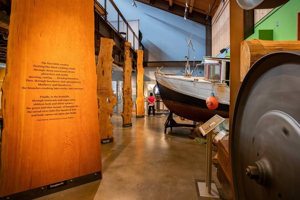 A museum exhibit with a wooden column displaying text, a boat, and a large gear.