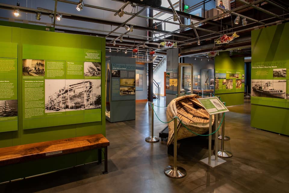 Uma exposição de museu com paredes verdes exibindo informações históricas sobre navegação marítima, um modelo de barco de madeira e um banco.