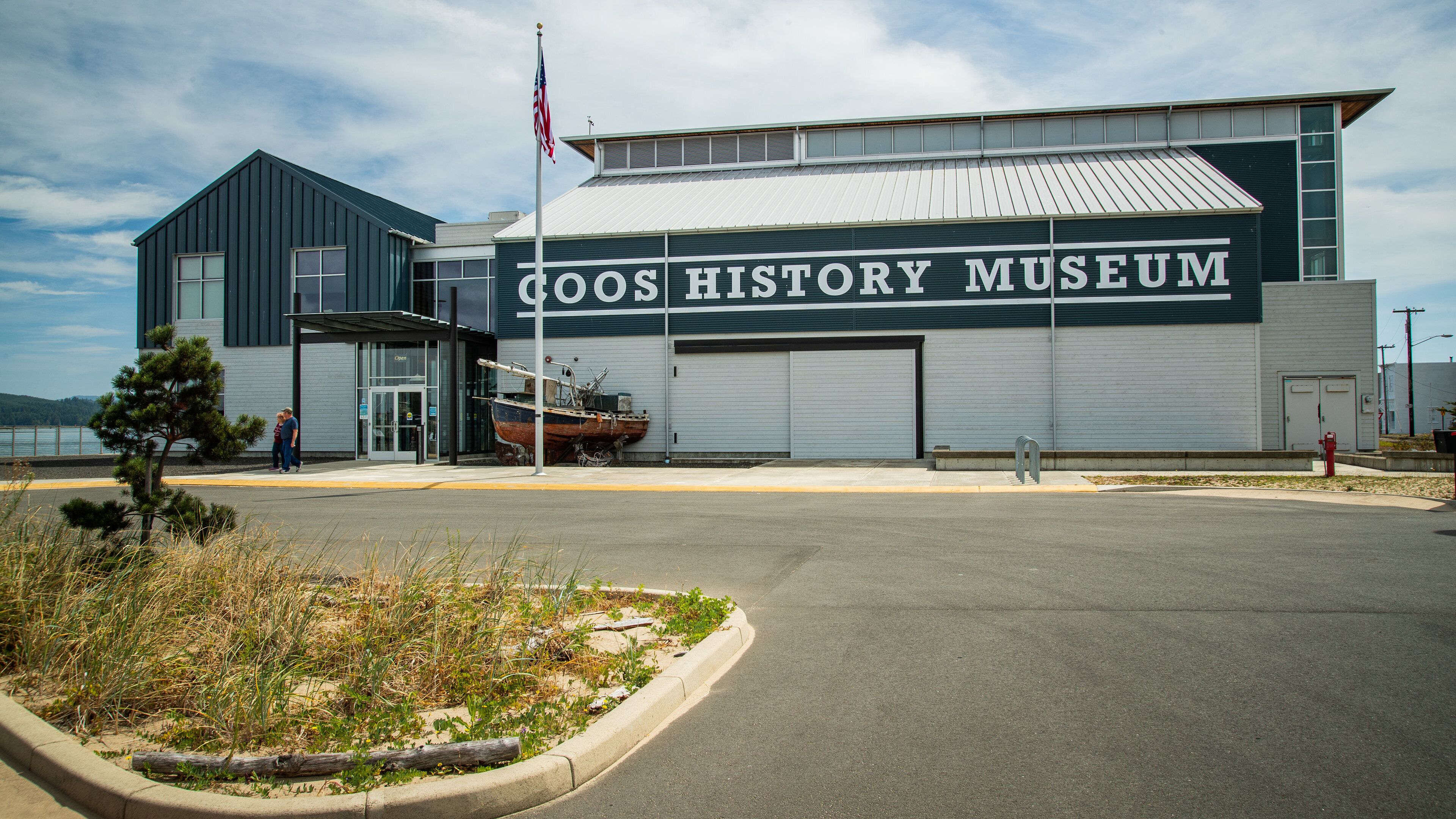 The exterior of the Coos History Museum with a large sign, a flag, and a boat displayed outside.