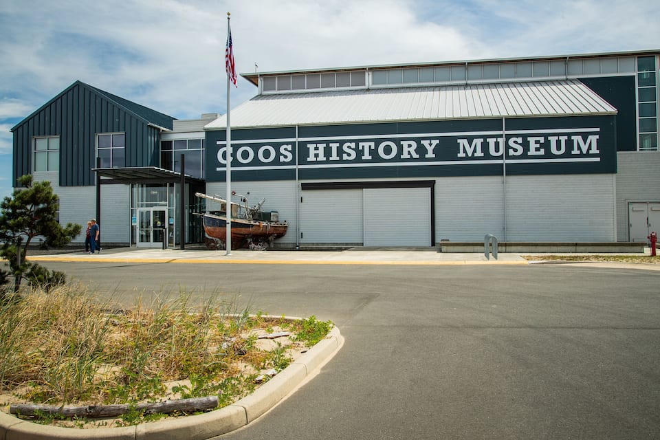 The exterior of the Coos History Museum with a large sign, a flag, and a boat displayed outside.