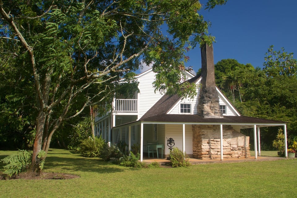 Una casa blanca con una chimenea de piedra, rodeada de exuberante vegetación y árboles.