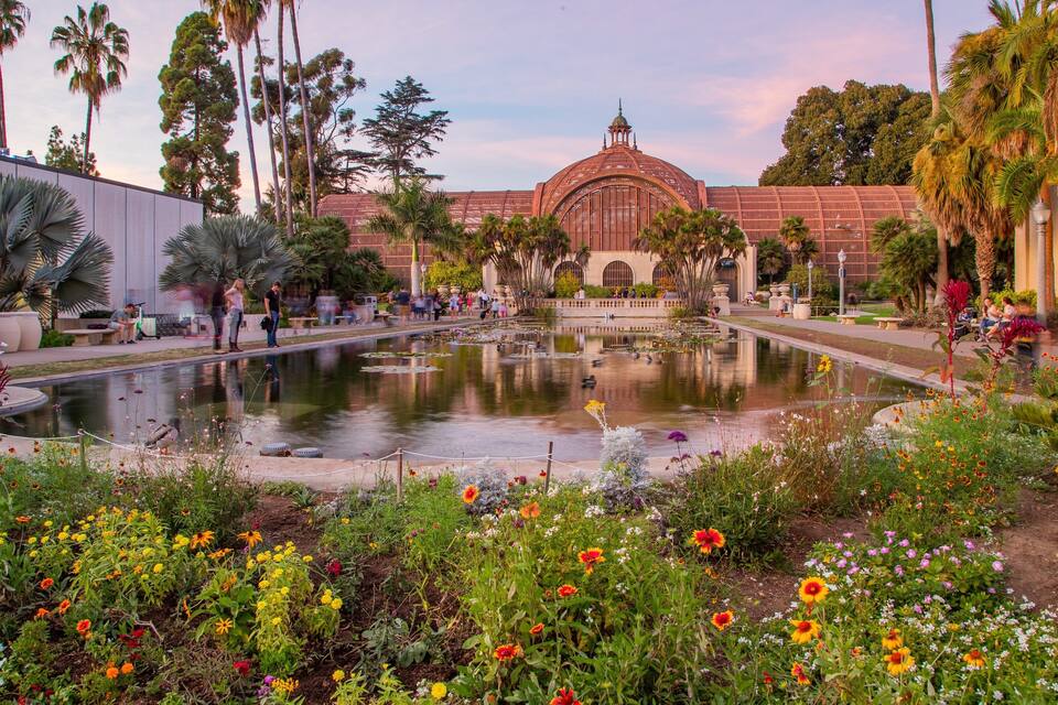 Un jardín con un estanque, flores coloridas, palmeras y un edificio grande con una cúpula.