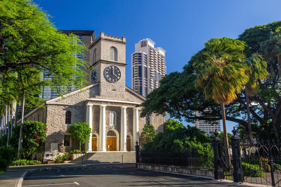 A historic stone building with a clock tower, surrounded by lush greenery and modern high-rise buildings.