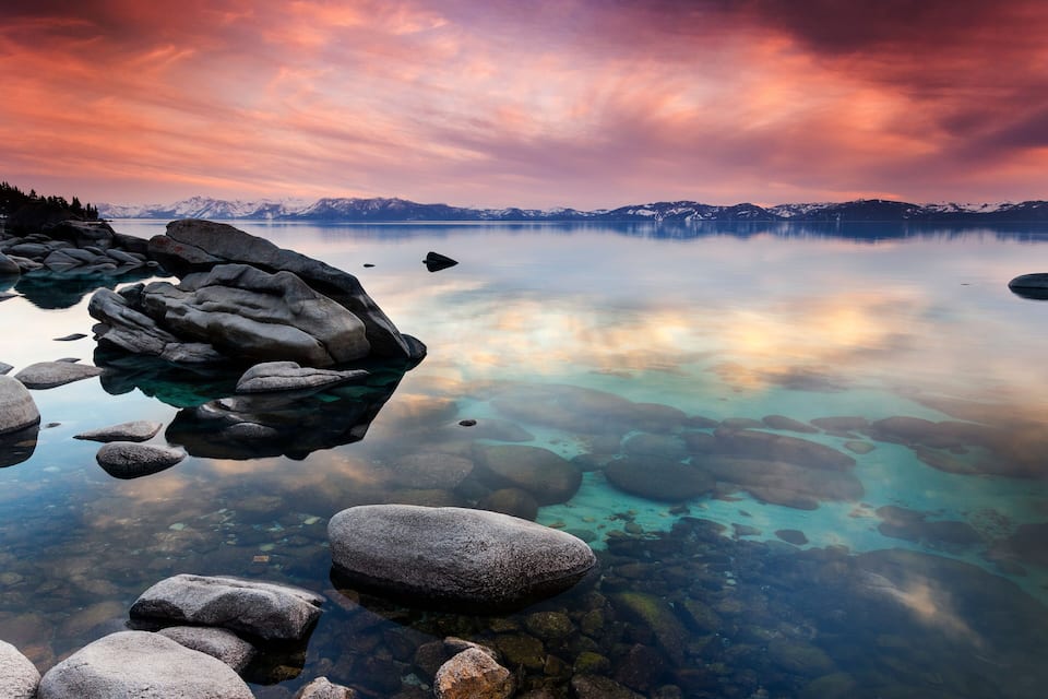 Un llac tranquil amb aigua cristal·lina que reflecteix el cel, envoltat de roques grans i muntanyes al fons.