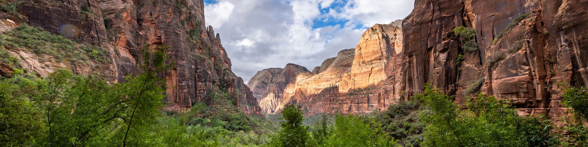A canyon with steep rock faces, dense green vegetation, and a partly cloudy sky.
