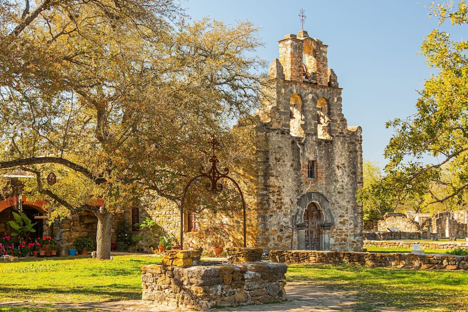 Una antigua torre de piedra con una cruz en la parte superior, rodeada de árboles y un muro de piedra.