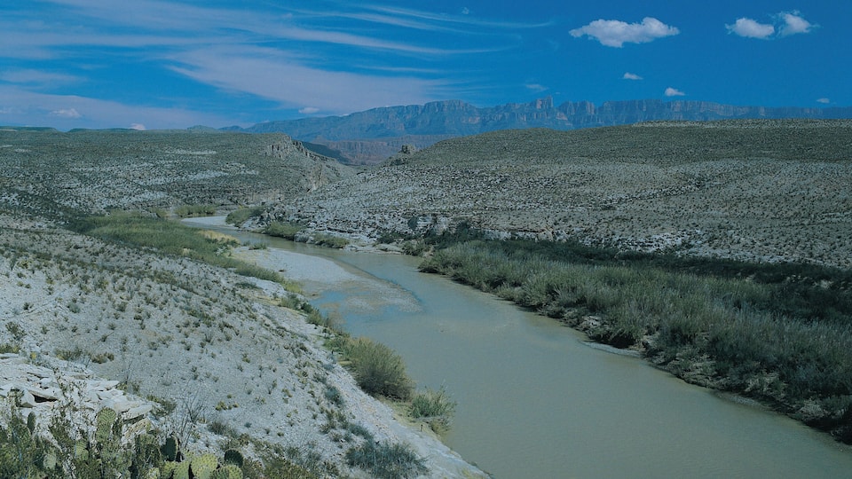 A river winding through a rocky desert landscape with sparse vegetation and distant mountains under a clear sky.