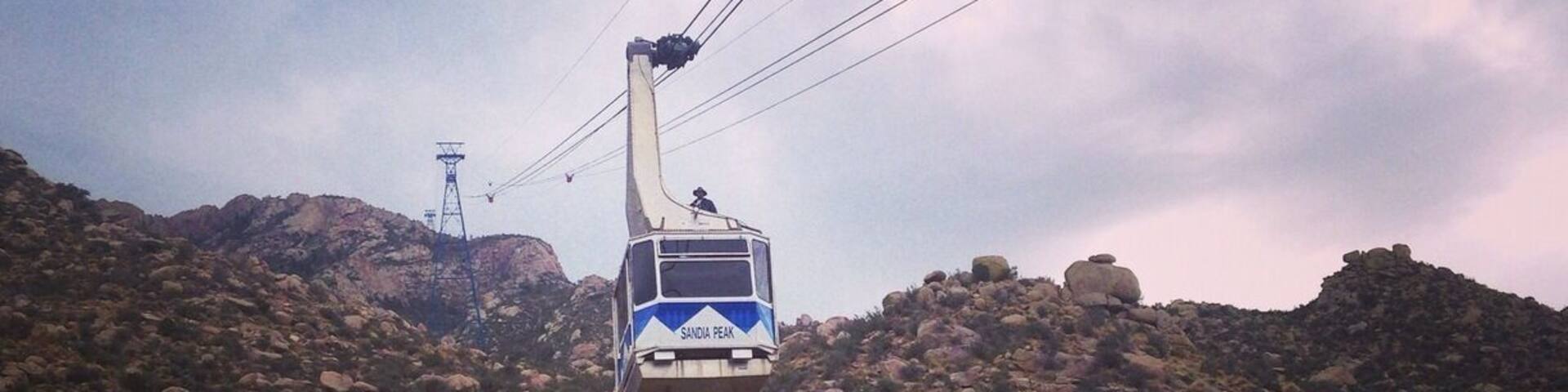 A cable car labeled 'SANDIA PEAK' moving over a rugged, rocky landscape with a sign indicating 'Cibola National Forest'.