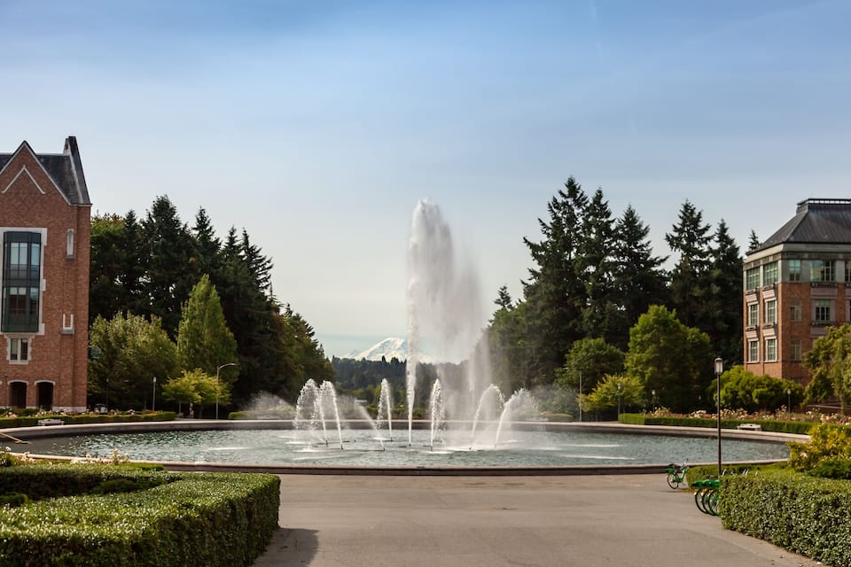 Une fontaine dotée de plusieurs jets d’eau, entourée d’un bassin circulaire, avec des bâtiments et des arbres en arrière-plan.