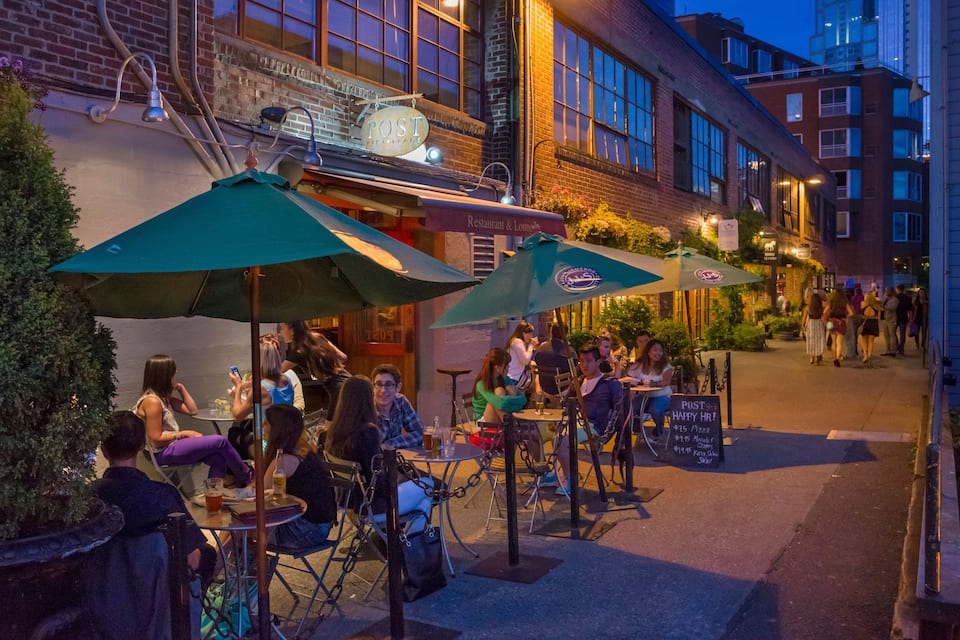 An outdoor dining area with tables and chairs under umbrellas, a brick building with a sign, and a sidewalk with people walking.