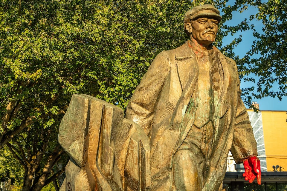 A statue of a man in a suit holding a book, set against a backdrop of green trees and a yellow building.