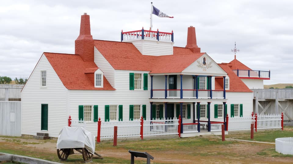 A white building with red roofs and green shutters, a flag, and a white picket fence.