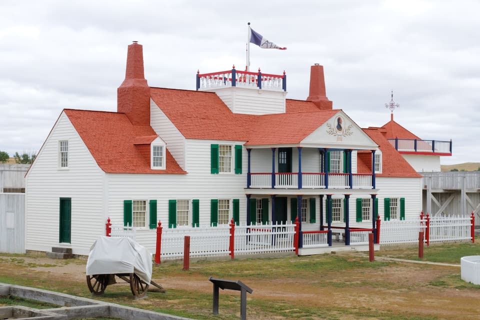 A white building with red roofs and green shutters, a flag, and a white picket fence.