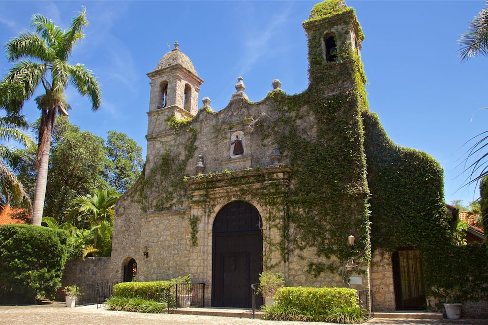 Une église historique en pierre, dotée d’un clocher, entourée d’une végétation luxuriante et de palmiers.