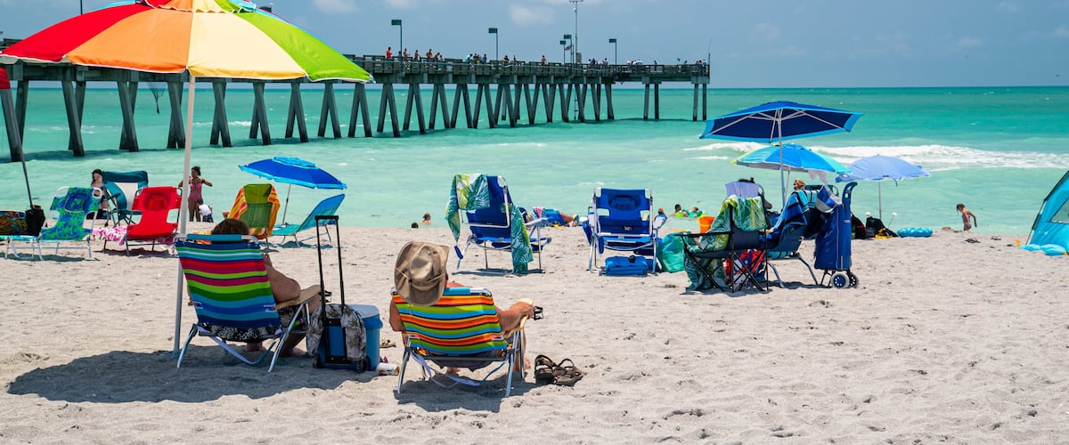 A beach with colorful umbrellas, lounge chairs, and a pier in the background.
