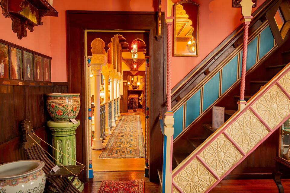 A grand staircase with ornate railings, a patterned carpet, and traditional decor including a large vase and a hanging lamp.