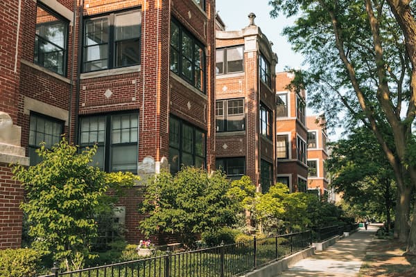 A row of red brick buildings with large windows, surrounded by green trees and a sidewalk.