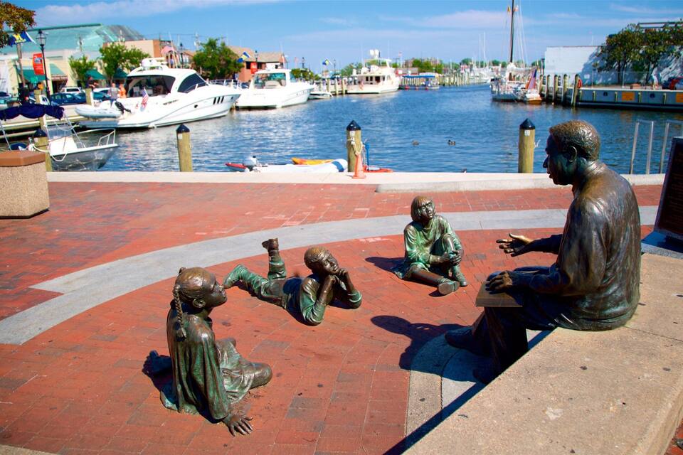 A waterfront with a statue of a man and children. Boats are docked in the background.