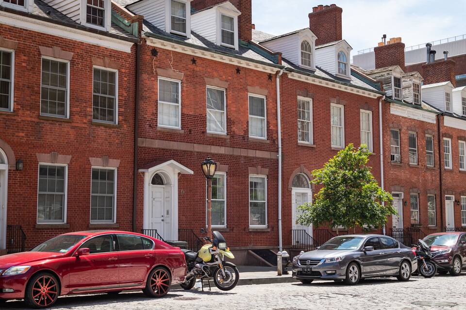 A red car parked on a cobblestone street in front of a red brick building with white trim and a tree in front.