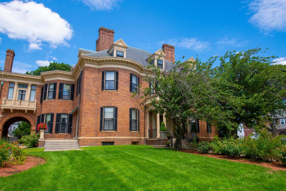 A large brick Victorian-style house with a well-manicured lawn and a tree in the foreground.