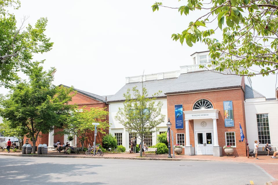 A building with a sign that reads "Whaling Museum" and a flag. There are trees, a bench, and a bicycle.