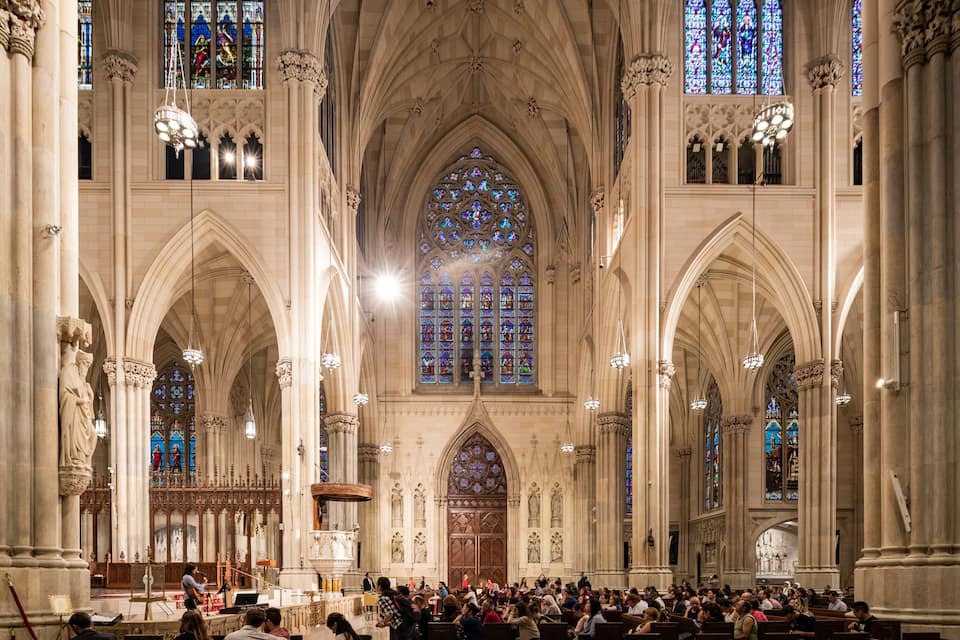 A grand cathedral interior with high vaulted ceilings, large stained glass windows, and ornate columns.