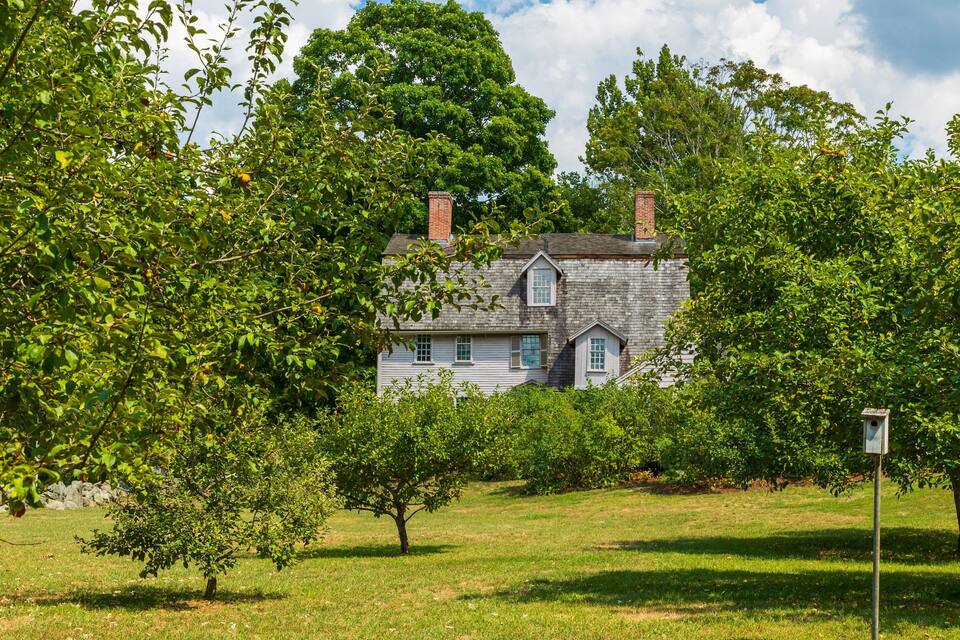 Une maison blanche avec une cheminée en briques, entourée d’arbres et d’herbe d’un vert luxuriant.