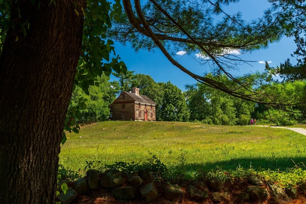 Une cabane en rondins historique, située dans un champ verdoyant, avec un mur de pierres au premier plan et un ciel bleu dégagé.