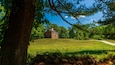 Une cabane en rondins historique, située dans un champ verdoyant, avec un mur de pierres au premier plan et un ciel bleu dégagé.