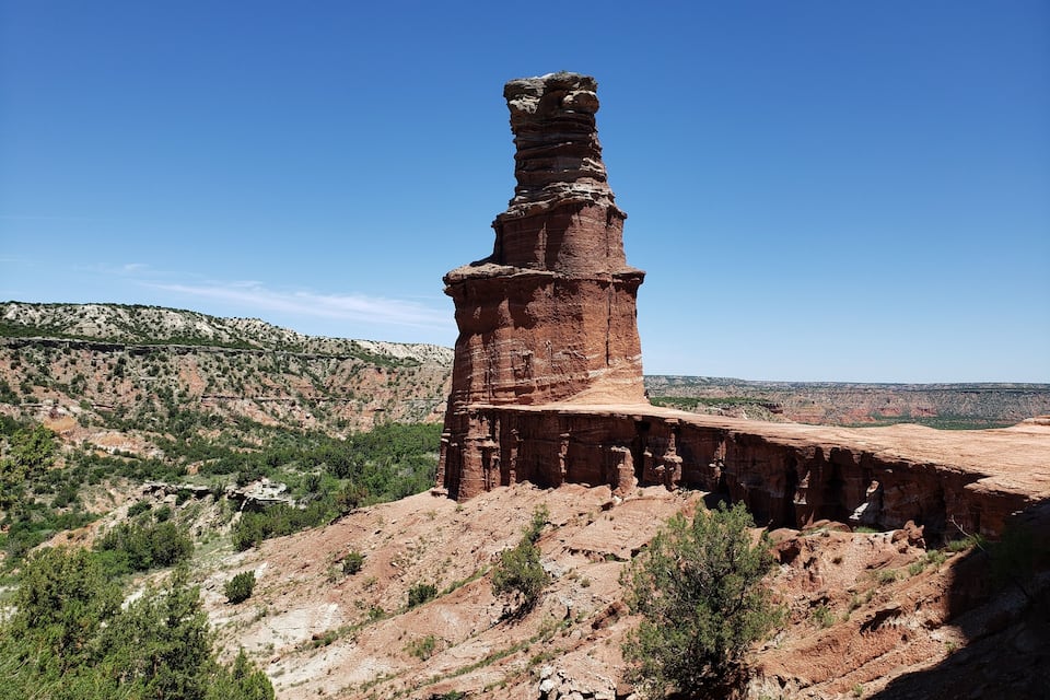 A tall, narrow rock formation with a flat top, surrounded by a rugged, hilly landscape with sparse vegetation.