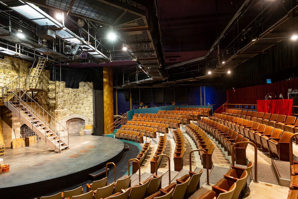 Teatre interior amb escenari, files de seients, mur de pedra i una escala.
