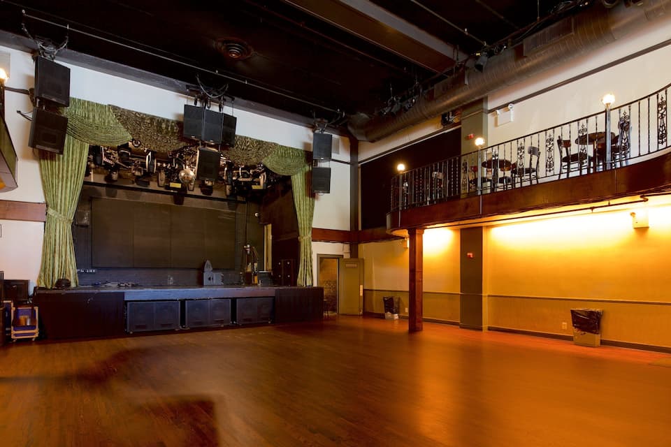 Ein geräumiger Saal mit Bühne, Holzboden und einem Balkon mit Metallgeländern.
