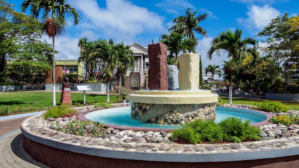 A circular fountain with a stone base, surrounded by a landscaped garden with palm trees and a building in the background.