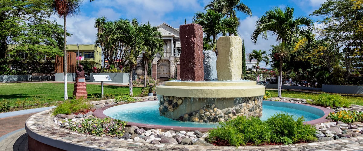A circular fountain with a stone base, surrounded by a landscaped garden with palm trees and a building in the background.