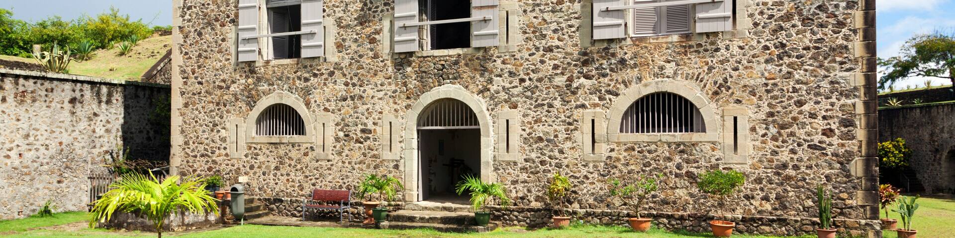 A stone building with arched windows and shutters, surrounded by a well-maintained lawn and potted plants.