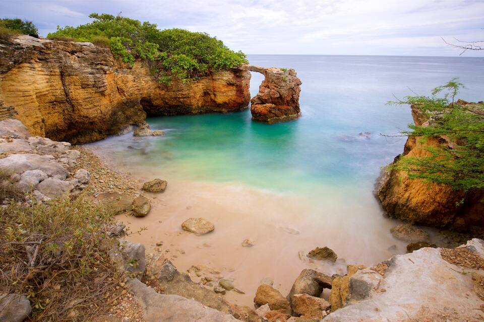 Paisagem costeira com mar azul cristalino, falésias rochosas e uma praia arenosa.