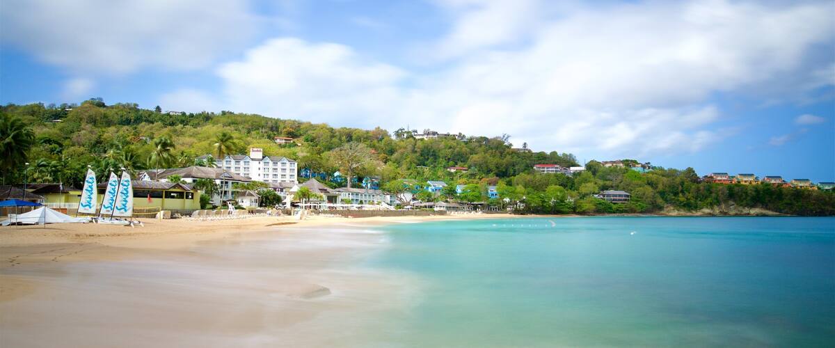 A beach with clear turquoise water, a sandy shore, and a backdrop of lush green hills with buildings.