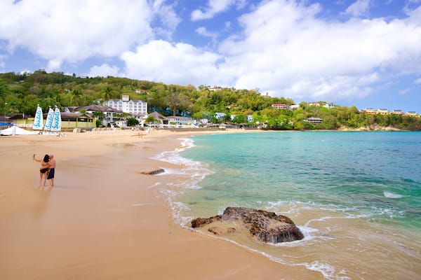 A beach with clear turquoise water, a rocky outcrop, and a backdrop of colorful buildings and lush greenery.
