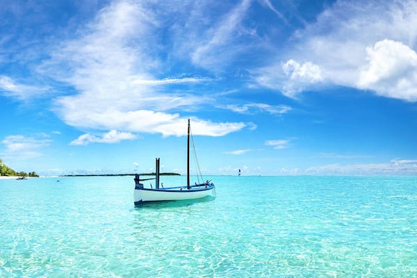 A clear turquoise sea with a small boat anchored near a sandy beach, palm trees, and a distant island.