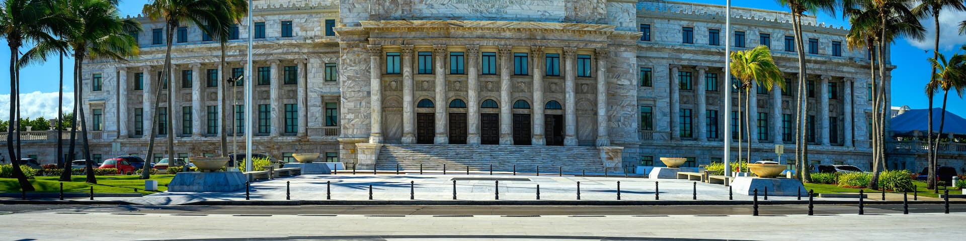 A large neoclassical building with a dome, two flags, palm trees, and a geometric plaza design.