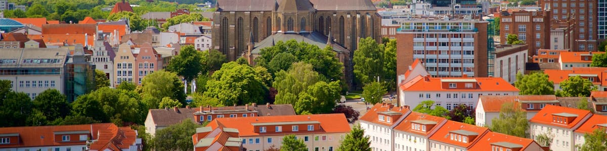 A historic church with a tall spire, surrounded by red-roofed buildings and lush green trees in a European city.