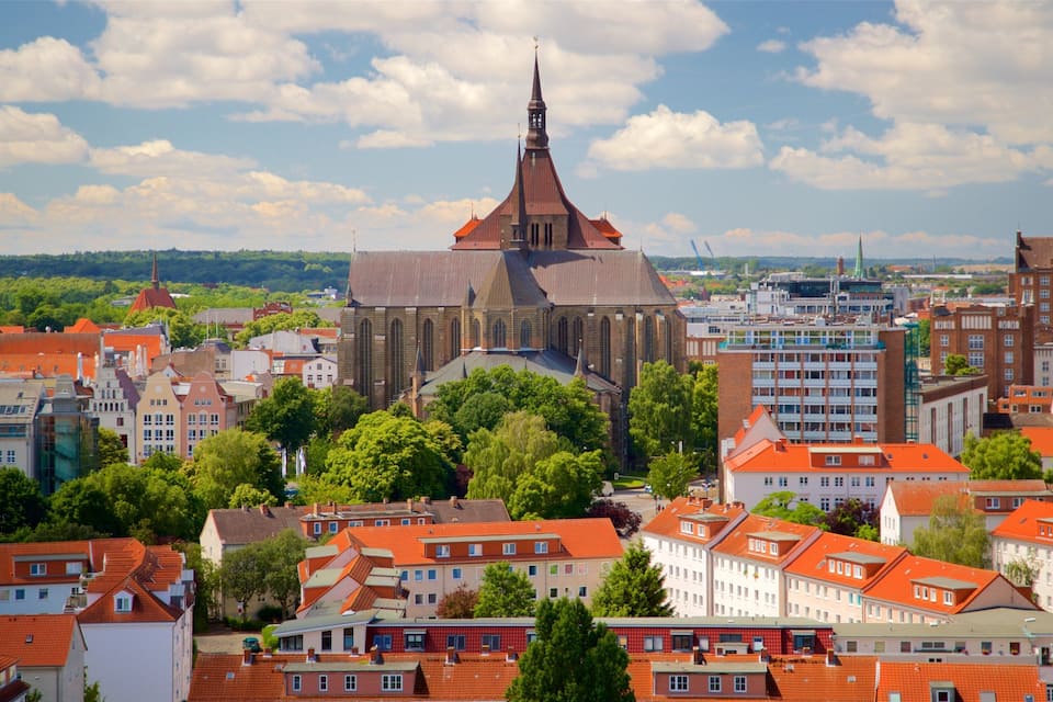 Eine historische Kirche mit einem hohen Turm, umgeben von Gebäuden mit roten Dächern und üppigen, grünen Bäumen in einer europäischen Stadt.