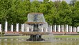 A multi-tiered fountain with water cascading down, surrounded by lush green trees and a building with white columns in the background.