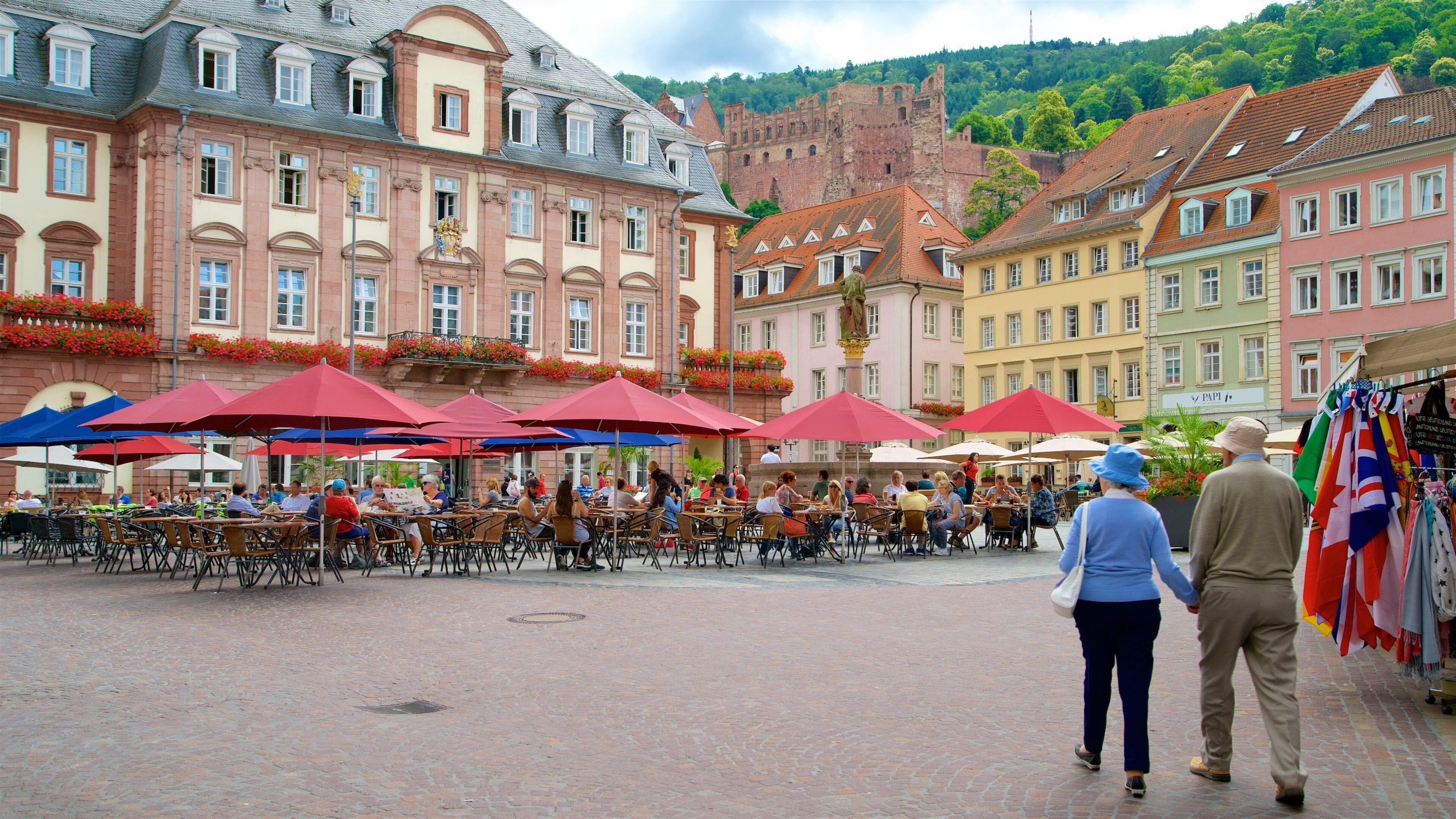 Ein Kopfsteinpflasterplatz mit Straßencafés, historischen Gebäuden und einer Burg im Hintergrund.