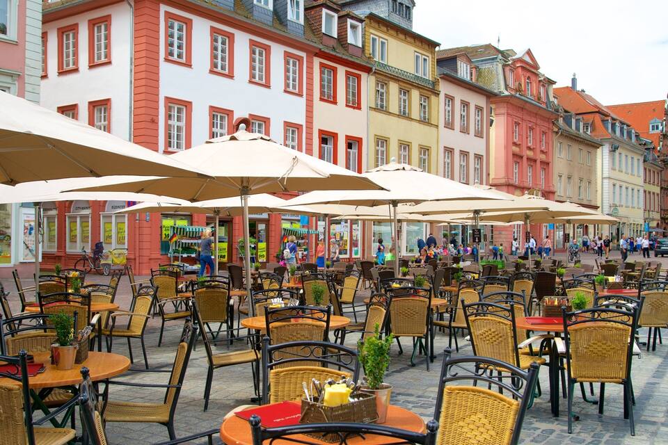 Um café agitado ao ar livre, com mesas e cadeiras de vime, cercado por edifícios coloridos e pedestres.