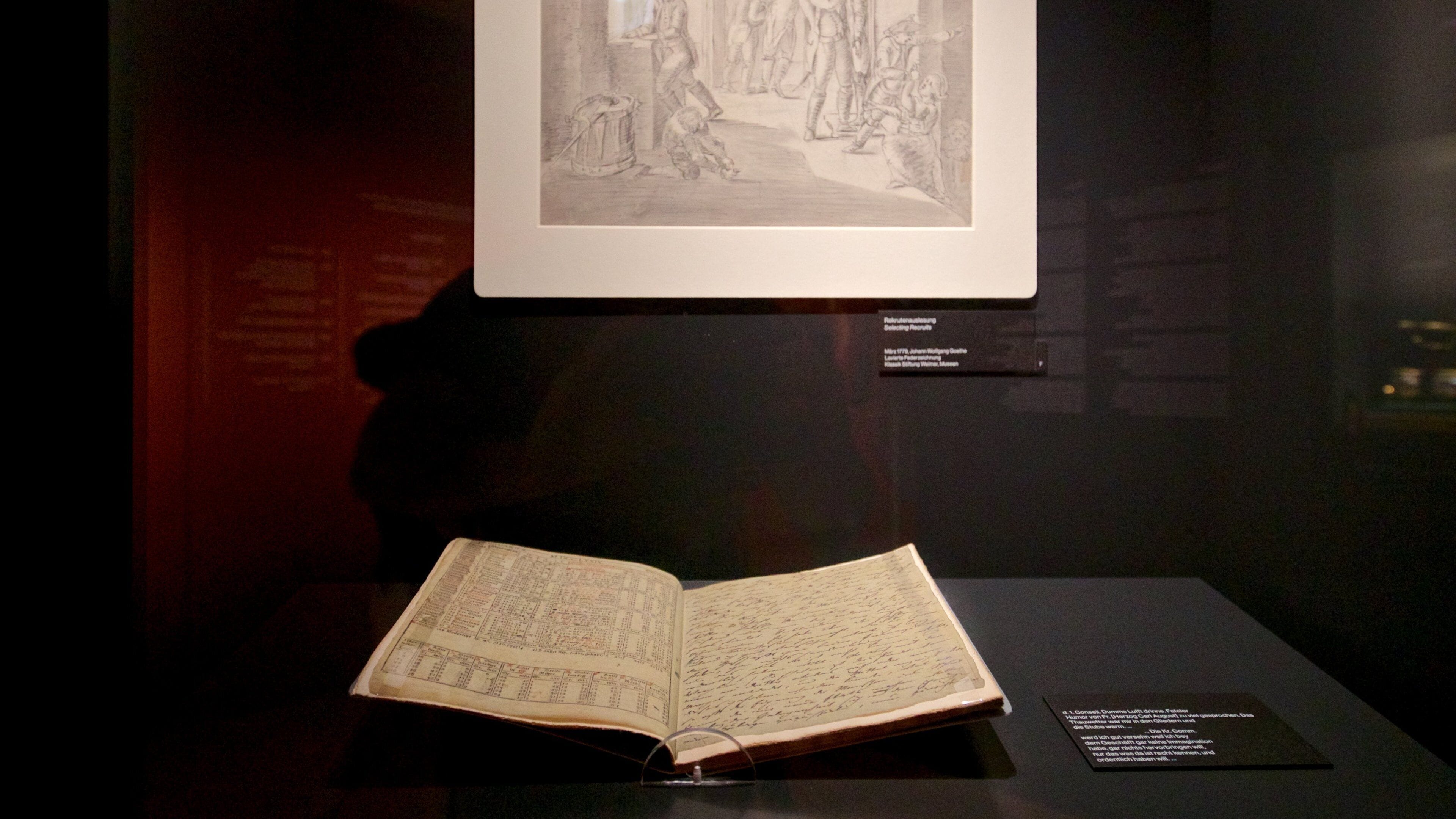 Un llibre obert amb notes manuscrites sobre una taula expositiva, un dibuix emmarcat a la paret i una petita placa informativa.