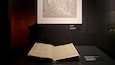 An open book with handwritten notes on a display table, a framed drawing on the wall, and a small informational plaque.